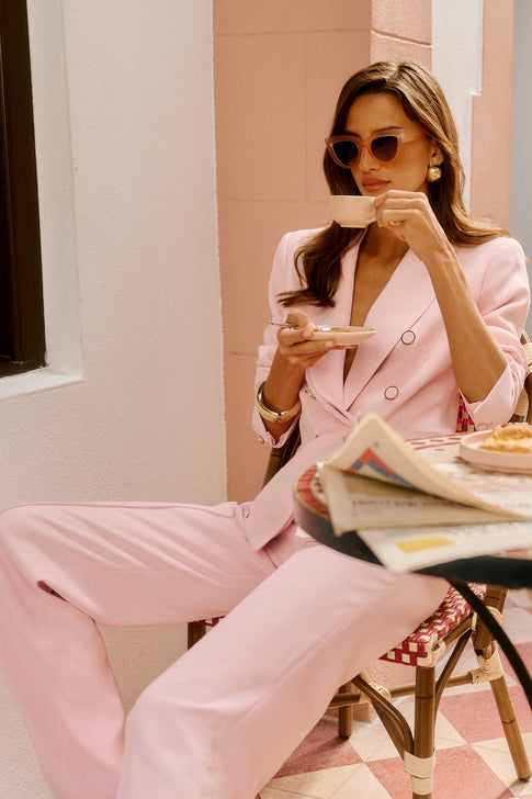 Woman in a pink suit sitting at a cafe, holding a cup and reading a newspaper.